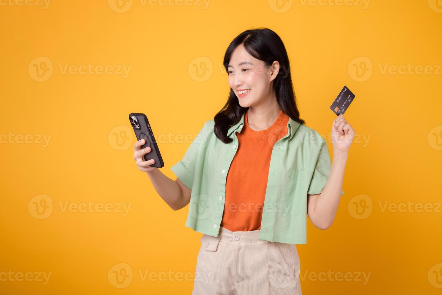 experiência a emoção do Móvel compras com uma vibrante jovem ásia mulher dentro dela 30s, vestindo laranja camisa e verde saltador, usando Smartphone enquanto apresentando crédito cartão em amarelo estúdio fundo. foto