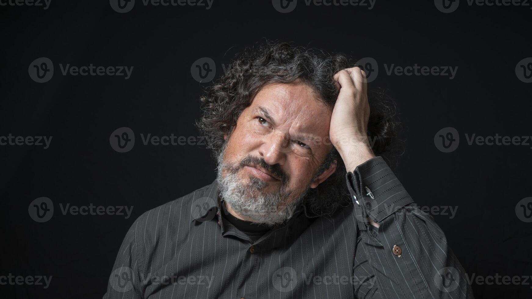 homem com branco barba e Preto encaracolado cabelo com confuso expressão, coçar cabeça com dele mão, vestindo Preto camisa contra Preto fundo foto