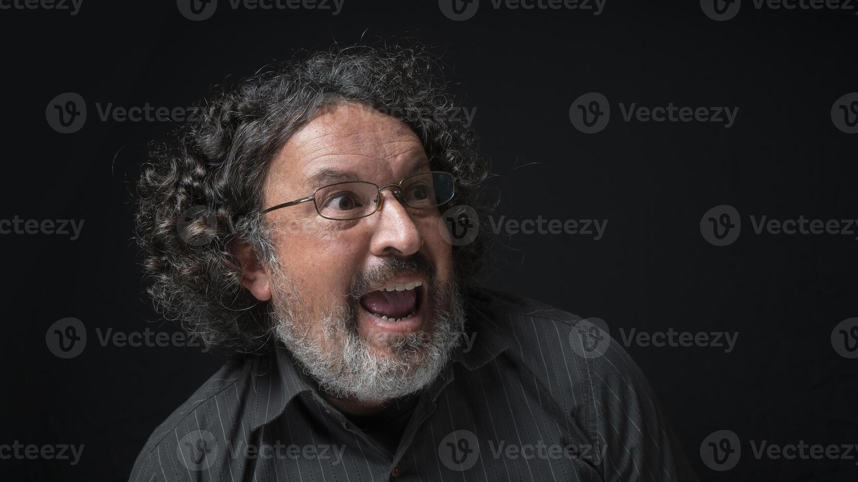 homem com branco barba e Preto encaracolado cabelo com expressão do surpresa e felicidade, olhando para a certo, vestindo Preto camisa contra Preto fundo foto