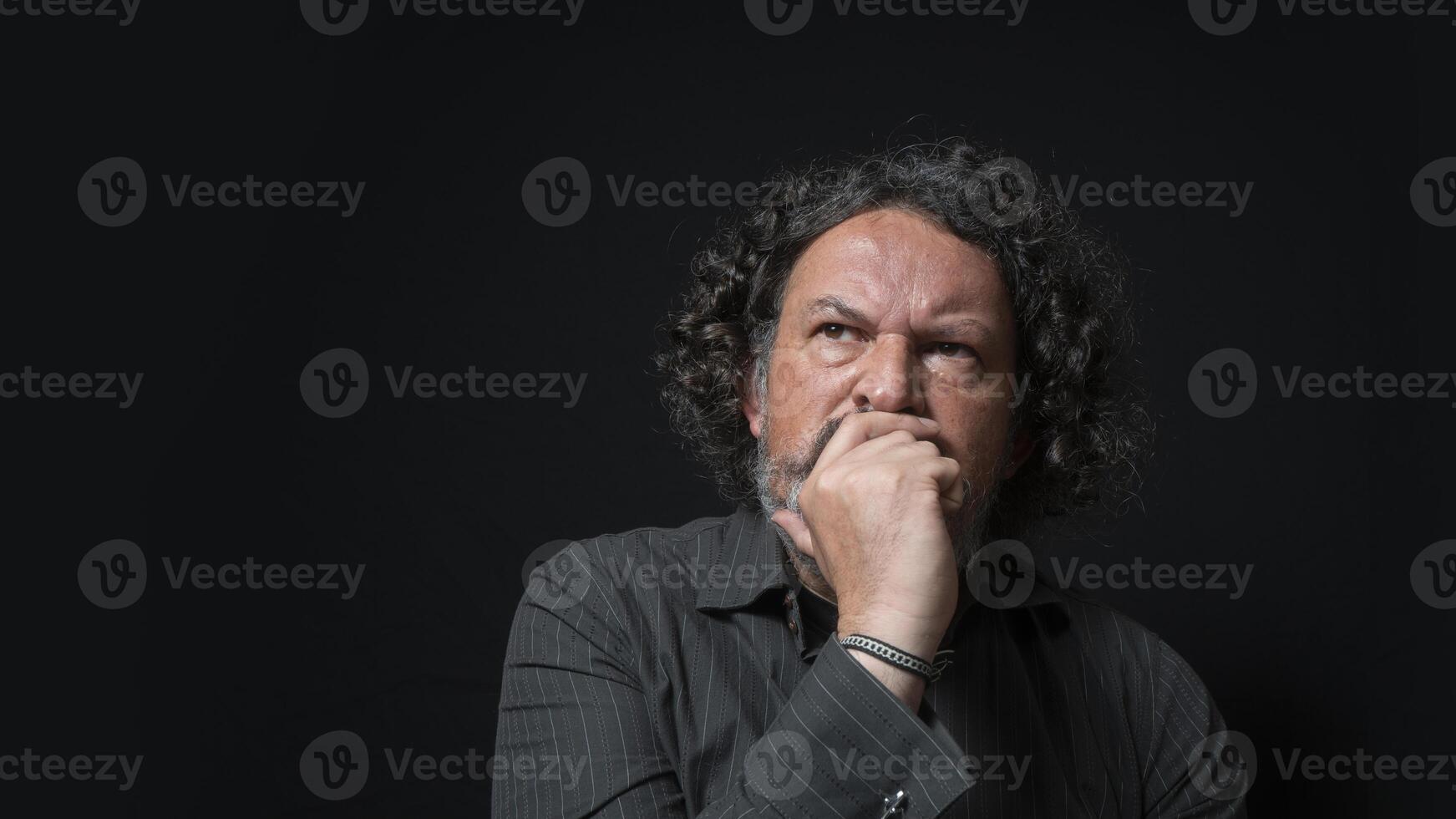 homem com branco barba e Preto encaracolado cabelo com duvidoso e Bravo expressão, com dele mão em dele face, vestindo Preto camisa contra Preto fundo foto