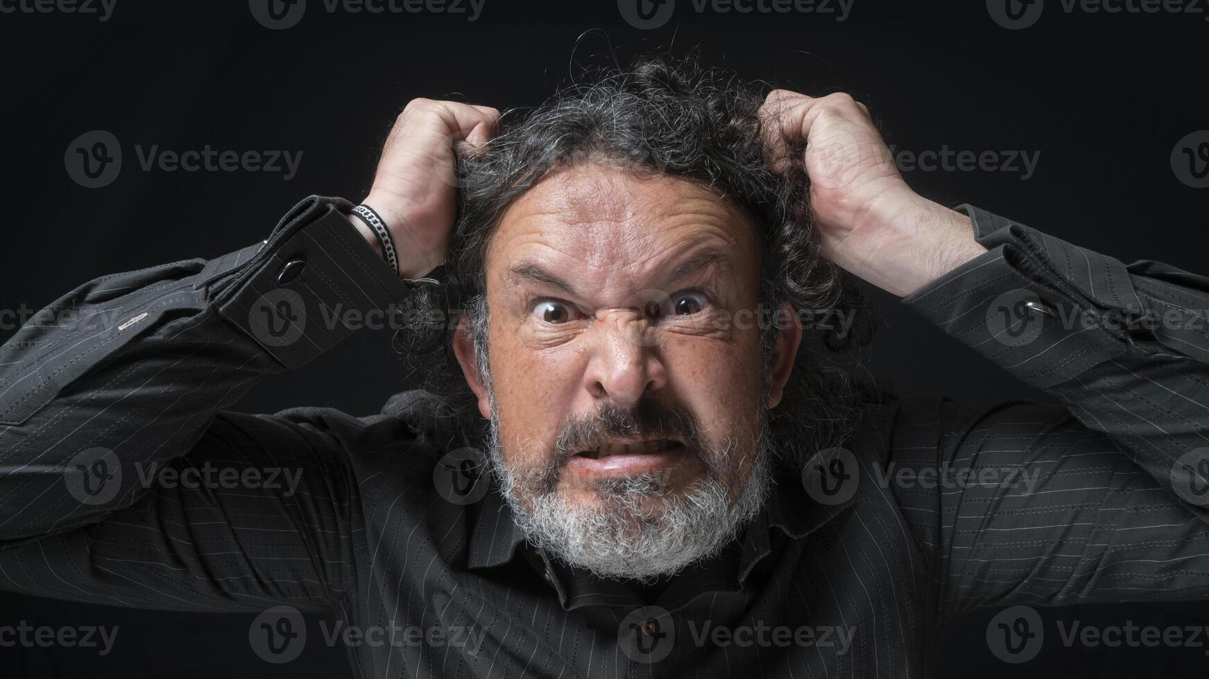 homem com branco barba e Preto encaracolado cabelo com muito Bravo expressão, agarrando dele cabelo com mãos, vestindo Preto camisa contra Preto fundo foto