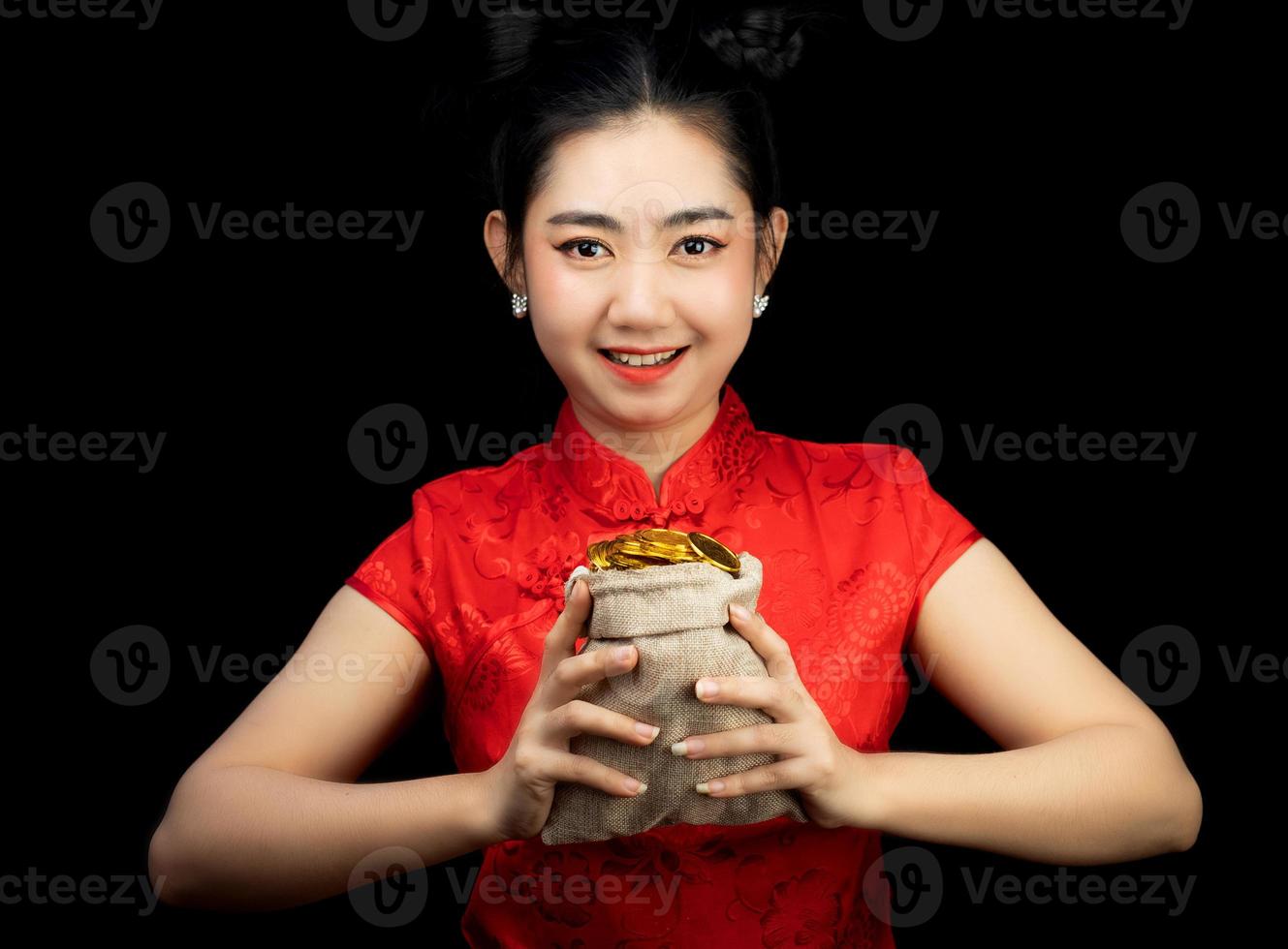 retrato asiático jovem, mulher vestido vermelho tradicional cheongsam segurando uma moeda de ouro em um saco no fundo preto foto