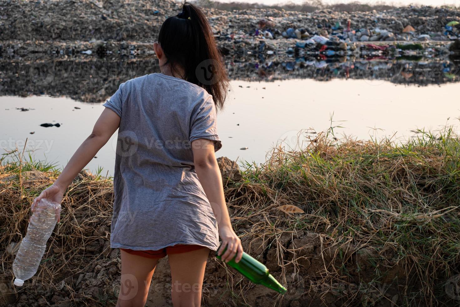 a mulher de volta está na mão com uma garrafa de vidro verde e uma garrafa de plástico transparente em uma montanha grande fundo de lixo foto