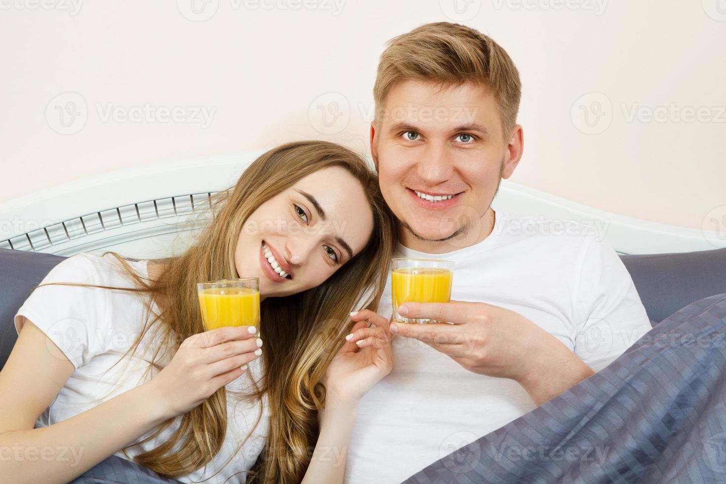 casal feliz bebendo suco de laranja enquanto estava deitado na cama no quarto pela manhã - estilo de vida saudável e conceito de nutrição foto