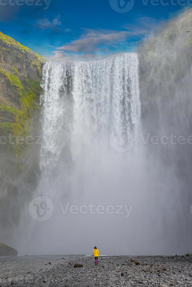 A poderosa e famosa cachoeira skogafoss com uma pessoa solitária em pé com uma jaqueta laranja, durante uma caminhada na Islândia, verão, vista panorâmica dramática em dia ensolarado e céu azul. foto