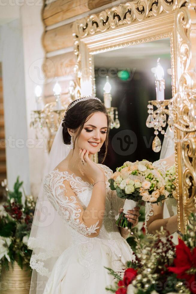 elegante morena noiva poses dentro uma branco vestir perto a espelho. retrato do a noiva, Casamento Maquiagem e Penteado, Casamento moda. lindo noiva dentro uma véu. foto