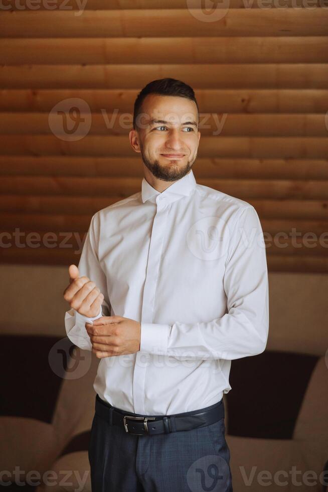 a noivo botões dele camisa de a janela. homem dentro uma camisa. masculino retrato. sessão de fotos às a janela. do noivo coleção. Casamento dia. bonito homem noivo abotoar dele camisa dentro uma hotel sala.