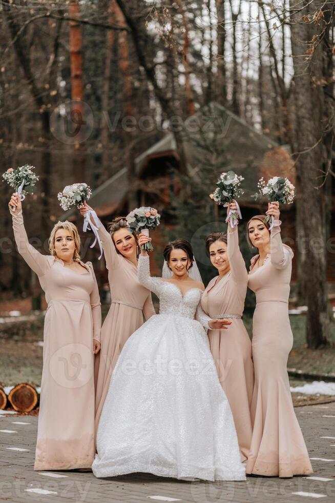 uma morena noiva dentro uma branco elegante vestir e dela amigos dentro cinzento vestidos pose com buquês. Casamento retrato dentro natureza, Casamento foto dentro luz cores.