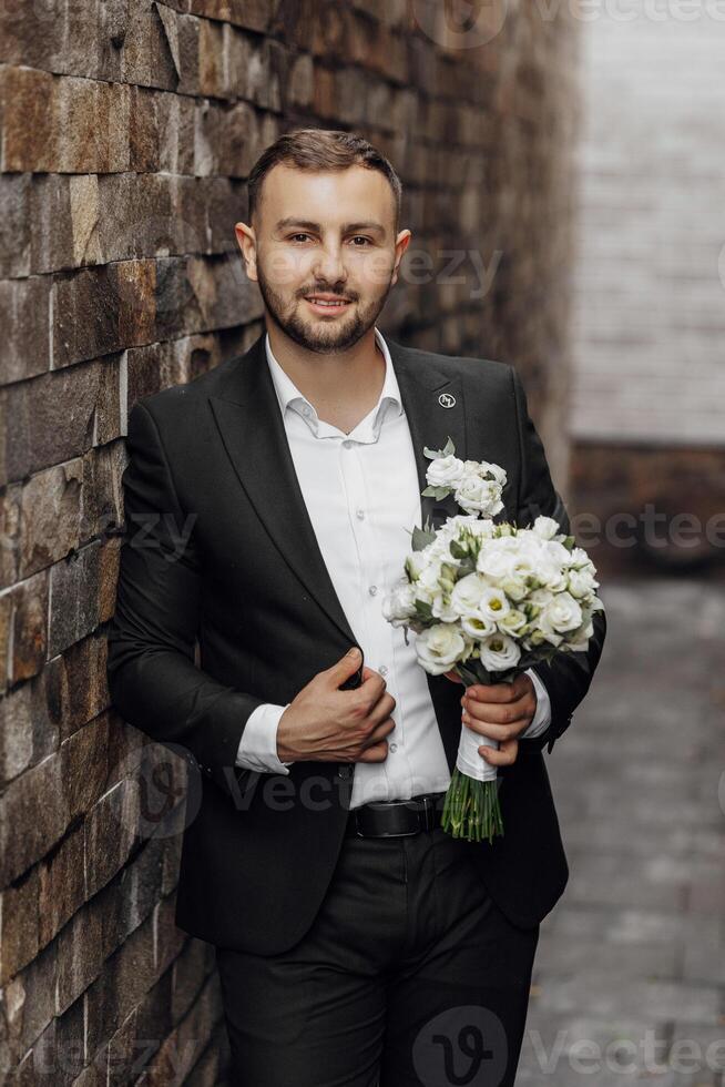 à moda e elegante jovem homem noivo dentro Preto terno e branco camisa com ramalhete do flores dentro mãos em tijolo fundo foto
