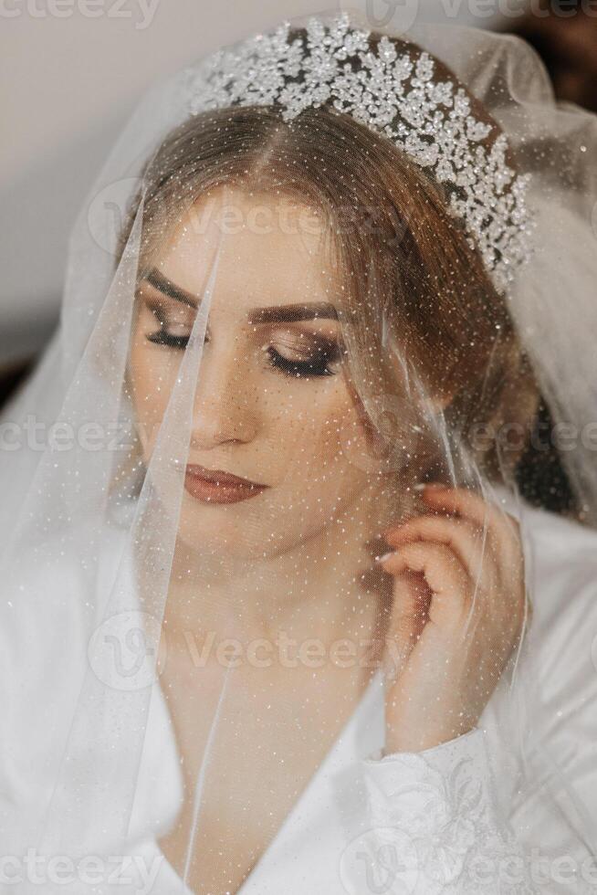 Casamento retrato. Loiras noiva dentro uma seda terno e tiara poses atrás uma puro véu enquanto sentado dentro uma cadeira dentro dela quarto foto