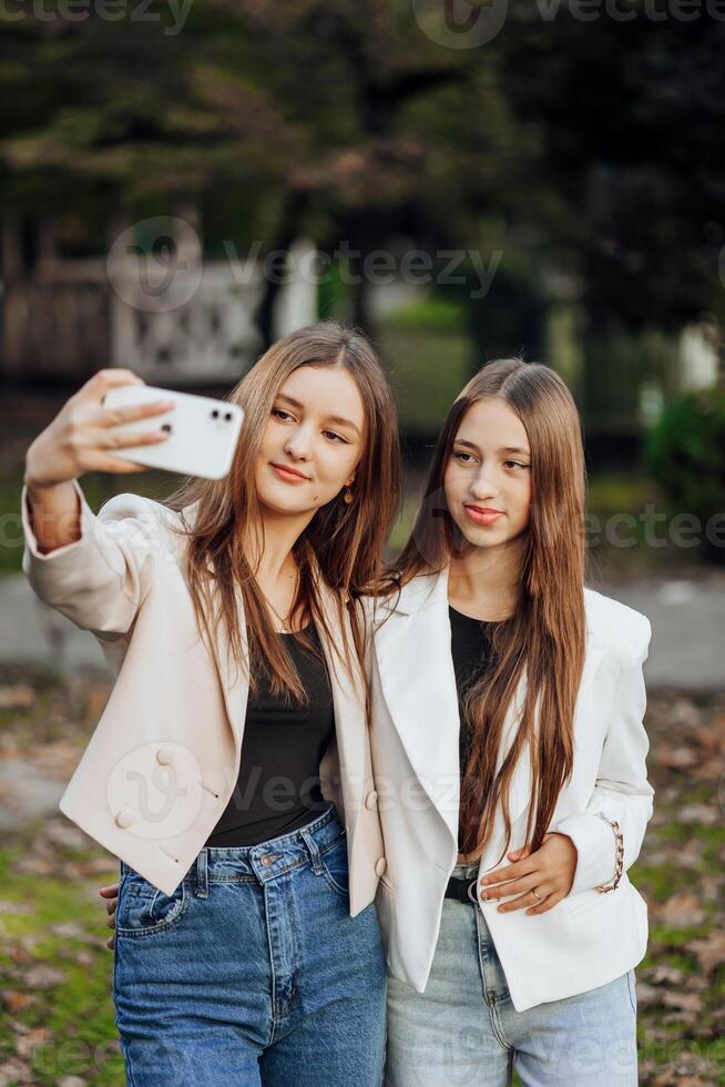 dois jovem mulheres levando uma selfie com uma inteligente Móvel telefone ao ar livre - feliz lindo fêmea amigos sorridente às a Câmera ao ar livre - estilo de vida conceito com alegre meninas desfrutando ao ar livre lazer. foto