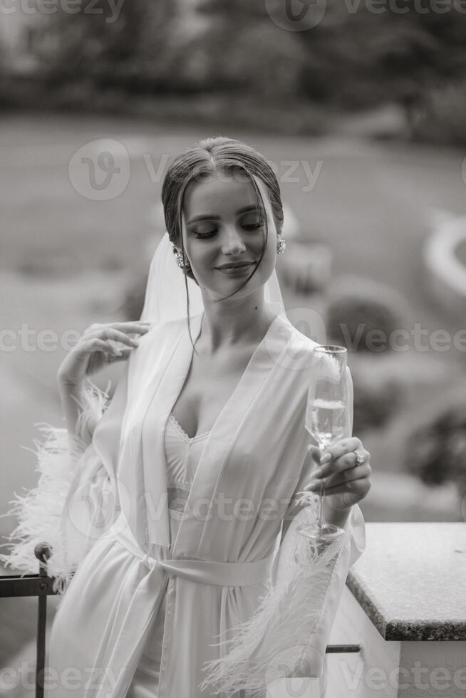 uma lindo noiva com uma grandes véu em dela cabelo, vestindo a elegante branco penhoar, em pé em uma sacada negligenciar a cidade, bebendo uma beber durante a Casamento manhã. Preto e branco foto