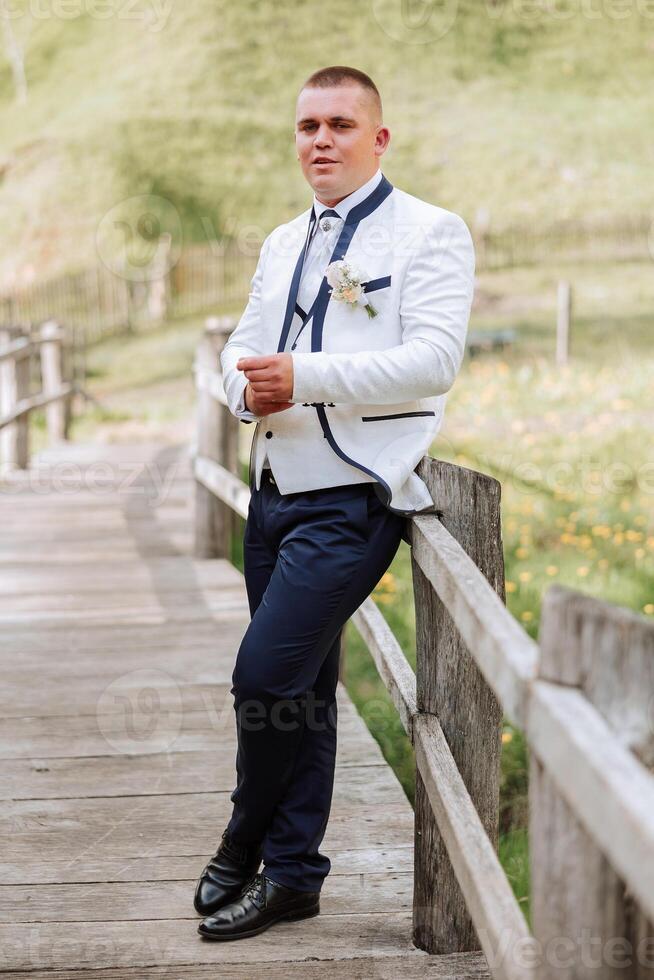 a noivo, dentro uma branco Jaqueta e Preto calça, poses inclinado em a grade e aperta uma botão em dele manga. Casamento retrato. foto