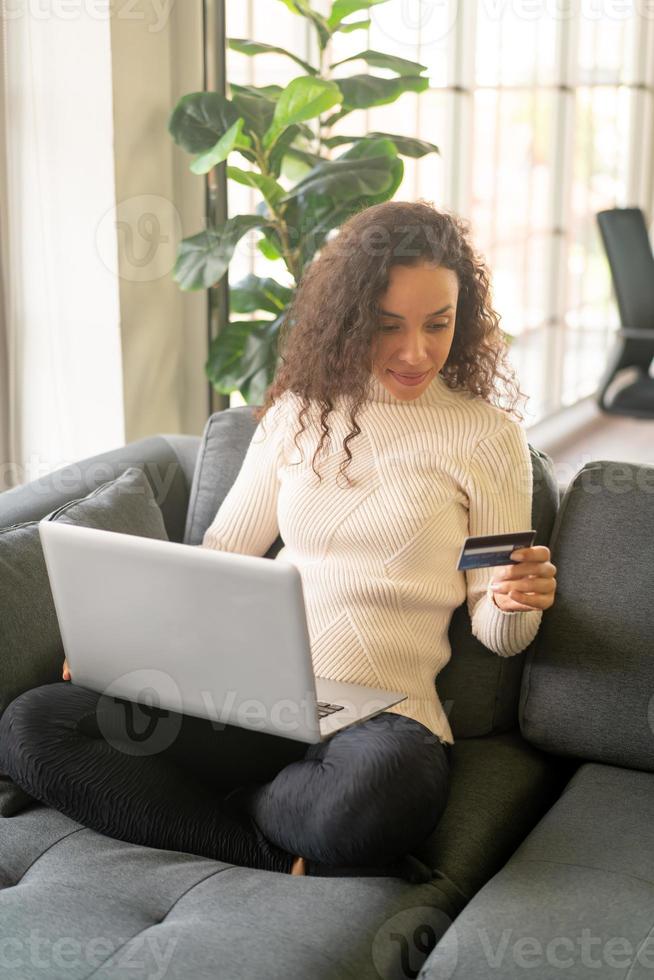 mulher latina usando laptop e segurando um cartão de crédito para fazer compras no sofá foto