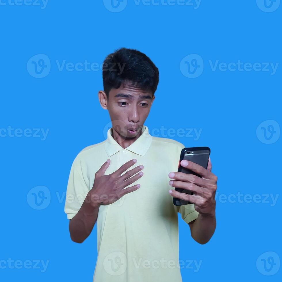 jovem ásia homem surpreso olhando às inteligente telefone vestindo amarelo camiseta, azul fundo. foto