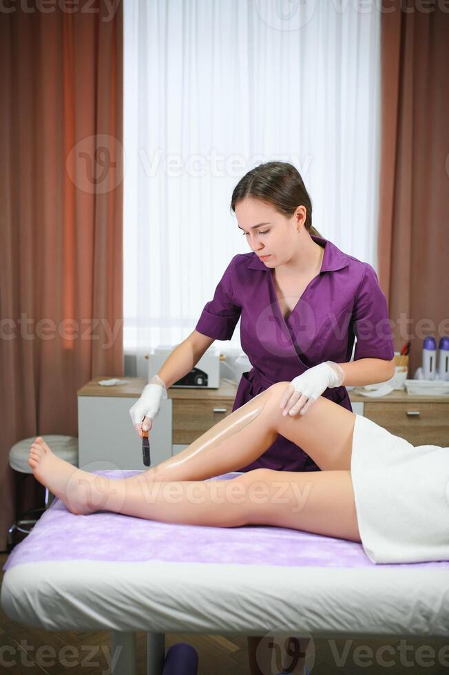 uma mestre aplica-se depilatório cera para uma jovem mulher perna para cabelo remoção. depilação com cera. beleza conceito. Lugar, colocar para texto. seletivo foco foto
