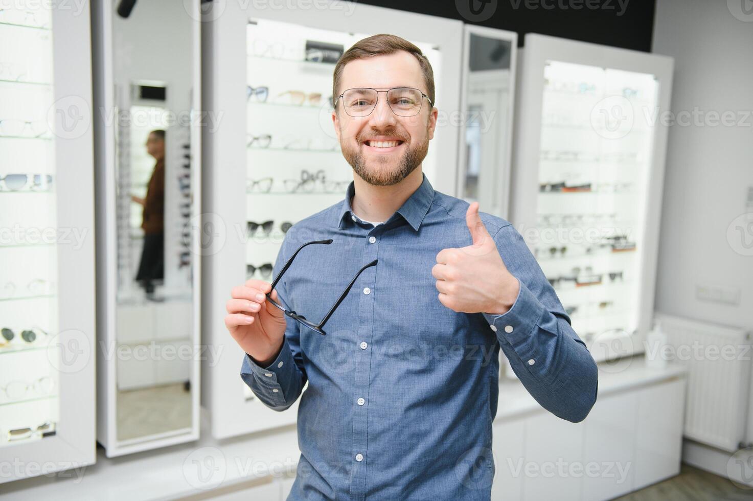 satisfeito cliente. Visão do feliz jovem masculino cliente vestindo Novo copos, em pé perto prateleira e mostruário com óculos. sorridente homem tentando em óculos foto