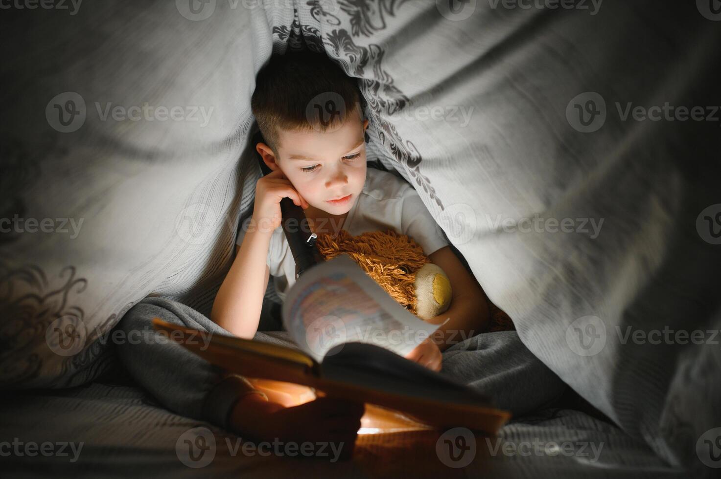 Garoto com lanterna lendo livro debaixo cobertor às lar. foto