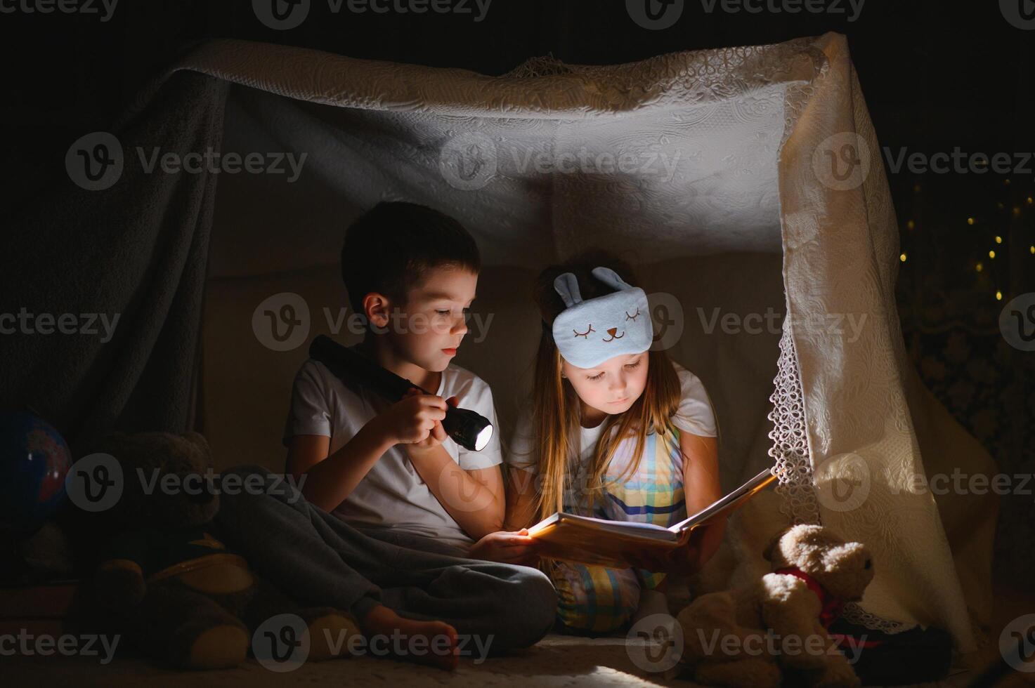 dois crianças com lanterna ler uma livro debaixo uma cobertor Como uma barraca. foto