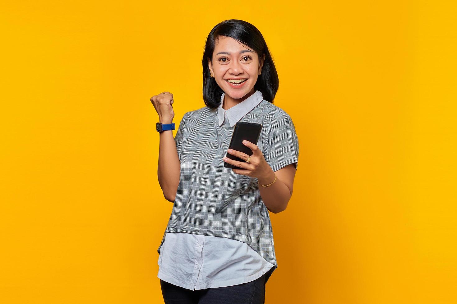 retrato de uma jovem asiática alegre segurando um telefone celular e celebrando o sucesso sobre o fundo amarelo foto