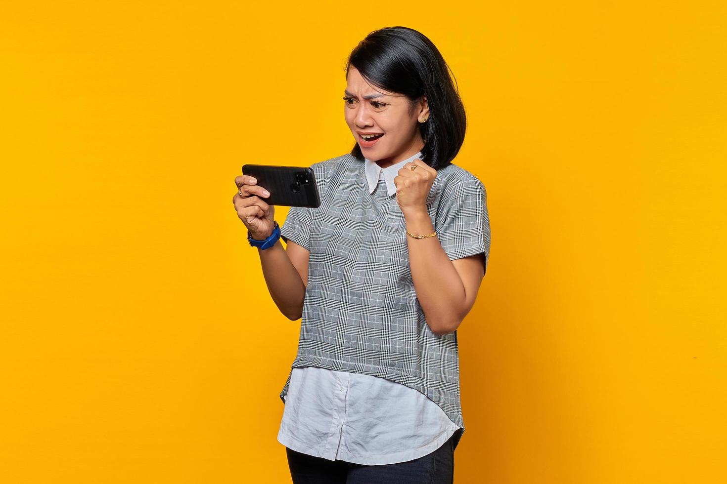 Retrato de uma jovem asiática alegre jogando videogame no celular e celebrando o sucesso no fundo amarelo foto