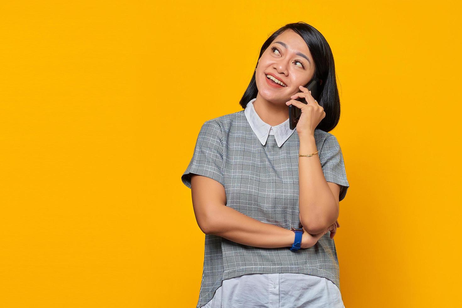 retrato de uma jovem mulher asiática sorridente, recebendo uma chamada no smartphone e olhando para o lado no fundo amarelo foto