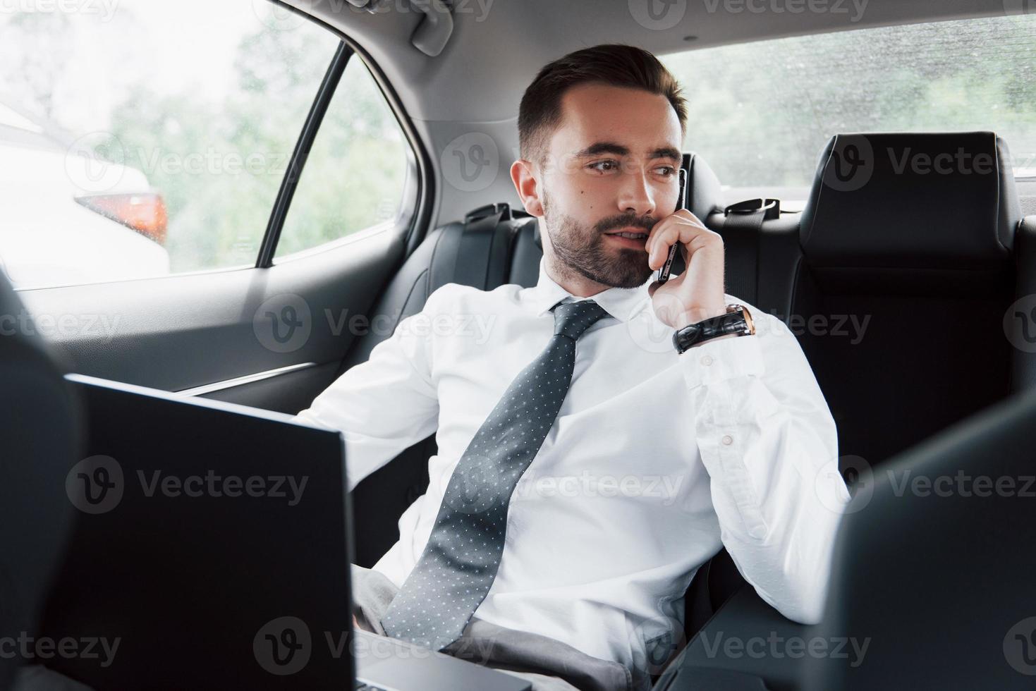 um jovem empresário trabalhando no laptop e falando ao telefone enquanto está sentado na parte de trás do carro. trabalha em movimento, aprecia seu tempo foto