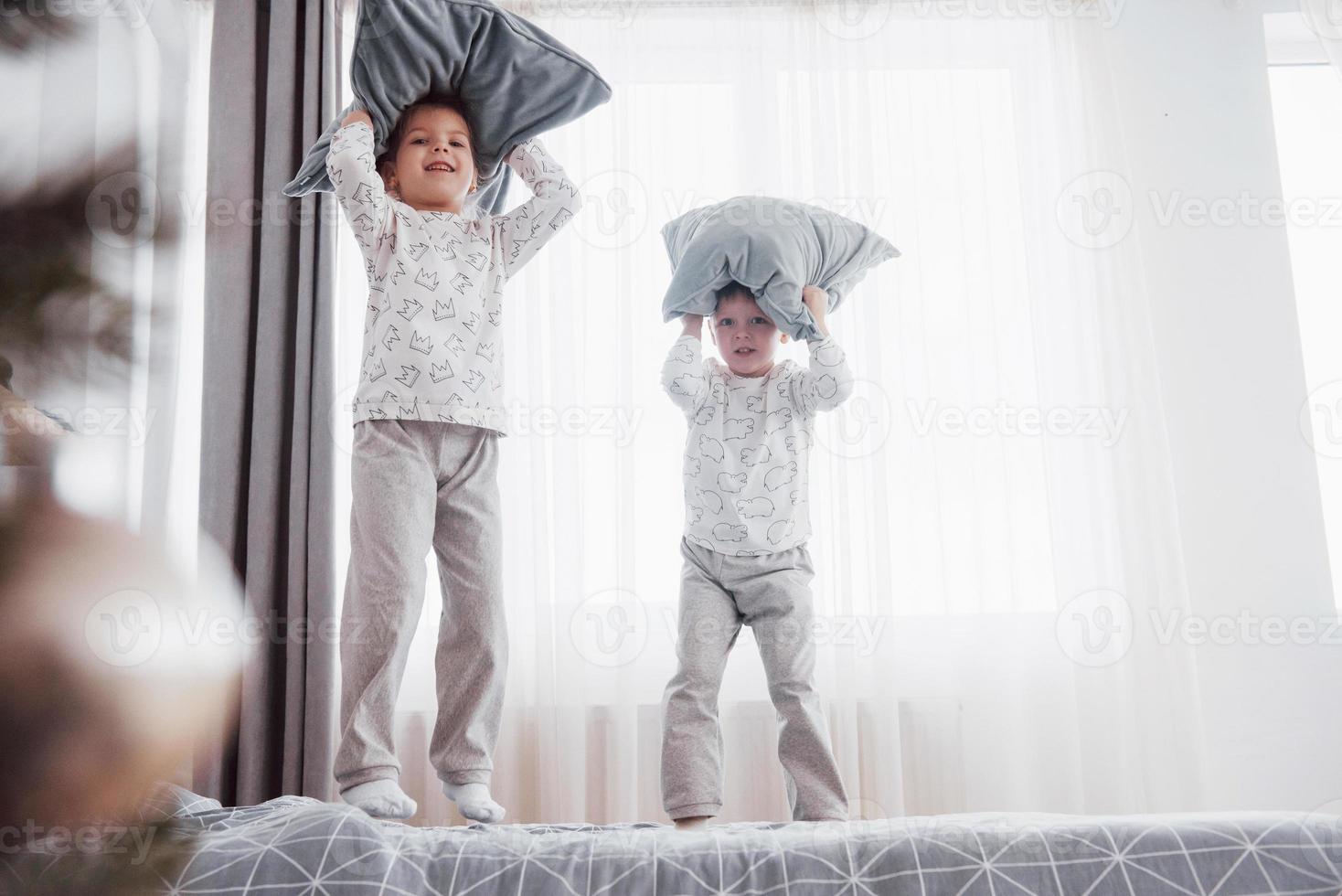 crianças brincando na cama dos pais. as crianças acordam em um quarto branco e ensolarado. menino e menina brincam de pijama combinando. manhã familiar foto