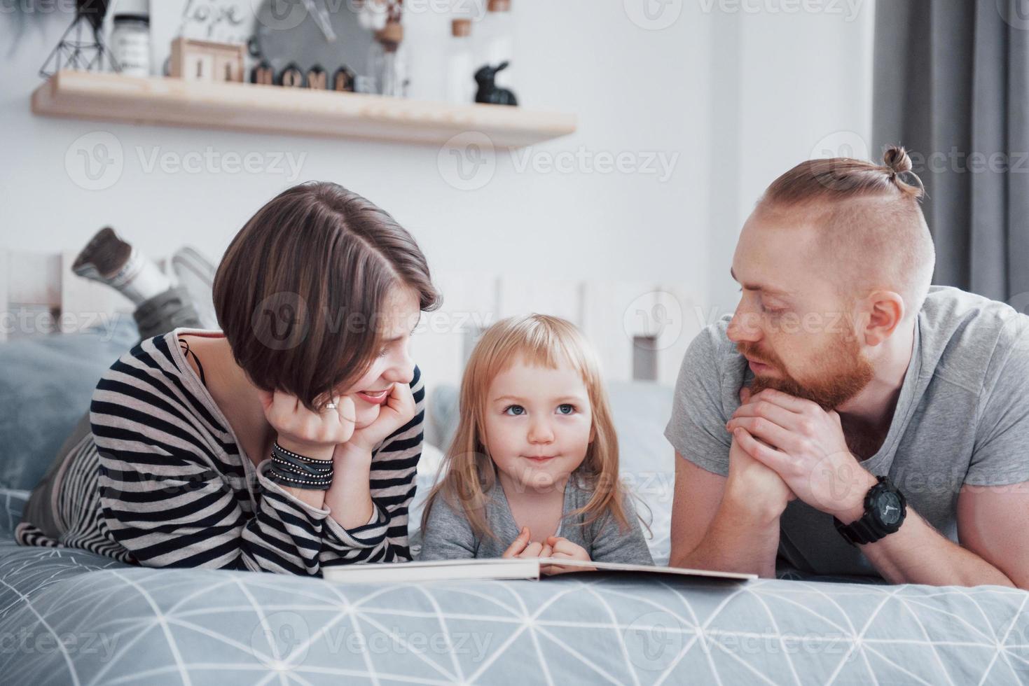 pai, mãe e filha lendo um livro infantil em um sofá na sala. família grande e feliz leu um livro interessante em um dia festivo. pais amam seus filhos foto