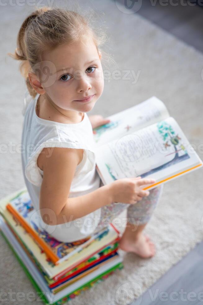 retrato do pequeno menina é sentado em pilha do crianças livros e folheando através uma livro com fada contos foto