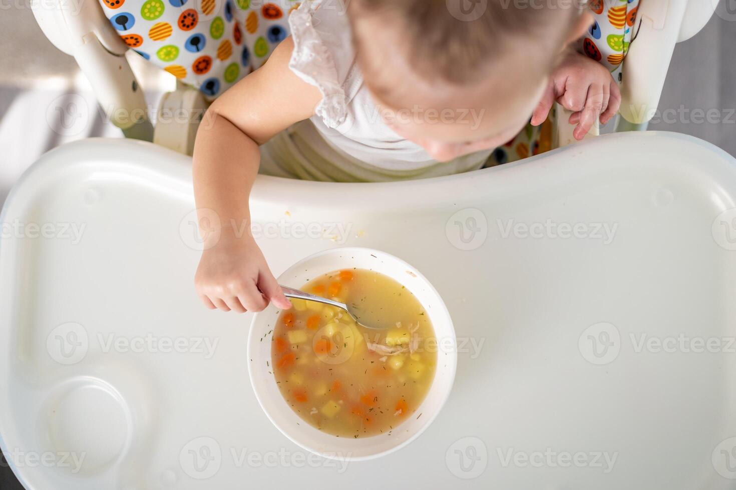 fofa bebê menina criança pequena sentado dentro a Alto cadeira e comendo dela almoço sopa às casa cozinha. foto