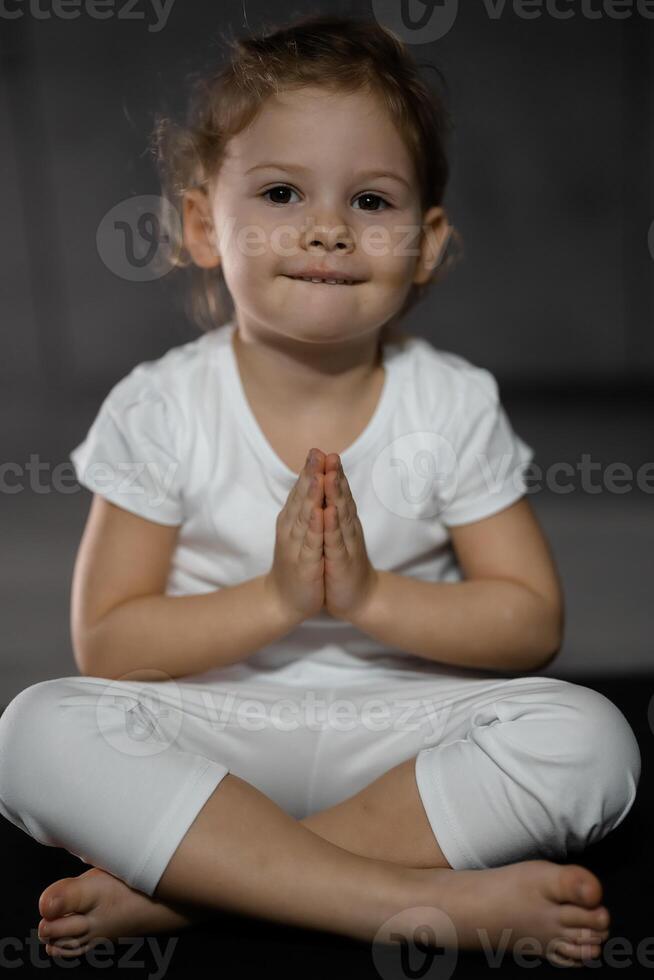 três anos velho pequeno menina meditando dentro uma lótus pose em uma cinzento fundo dentro Sombrio quarto foto