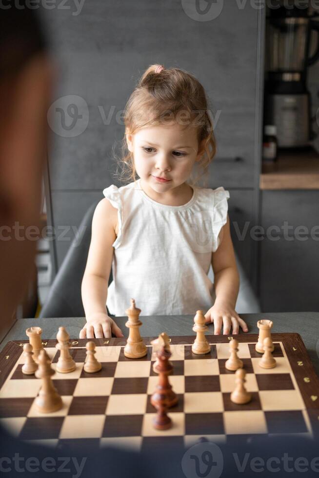 pequeno menina jogando xadrez com dela pai às a mesa dentro casa cozinha. a conceito cedo infância desenvolvimento e Educação. família lazer, comunicação e lazer. foto