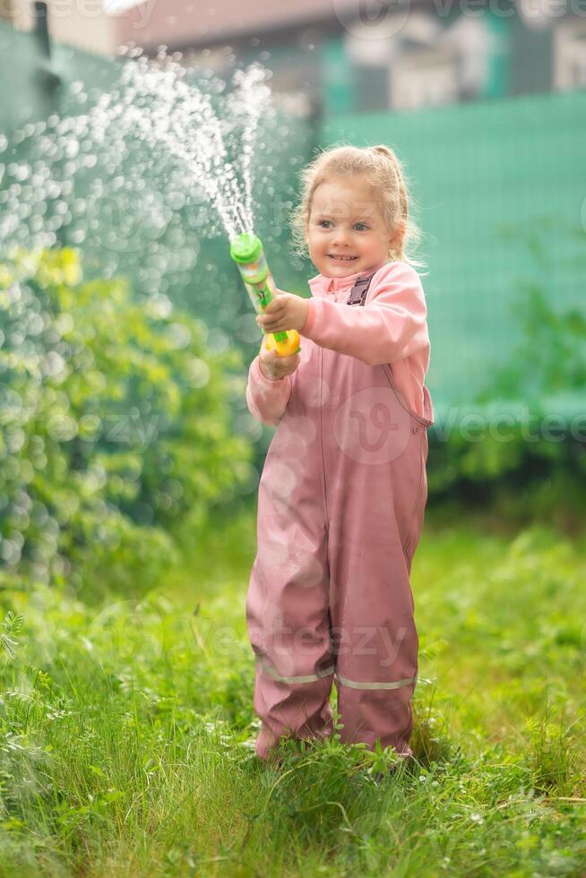 feliz rindo criança menina 2-3 ano velho vestindo à prova d'água roupas ter uma Diversão com água arma de fogo em casa quintal foto