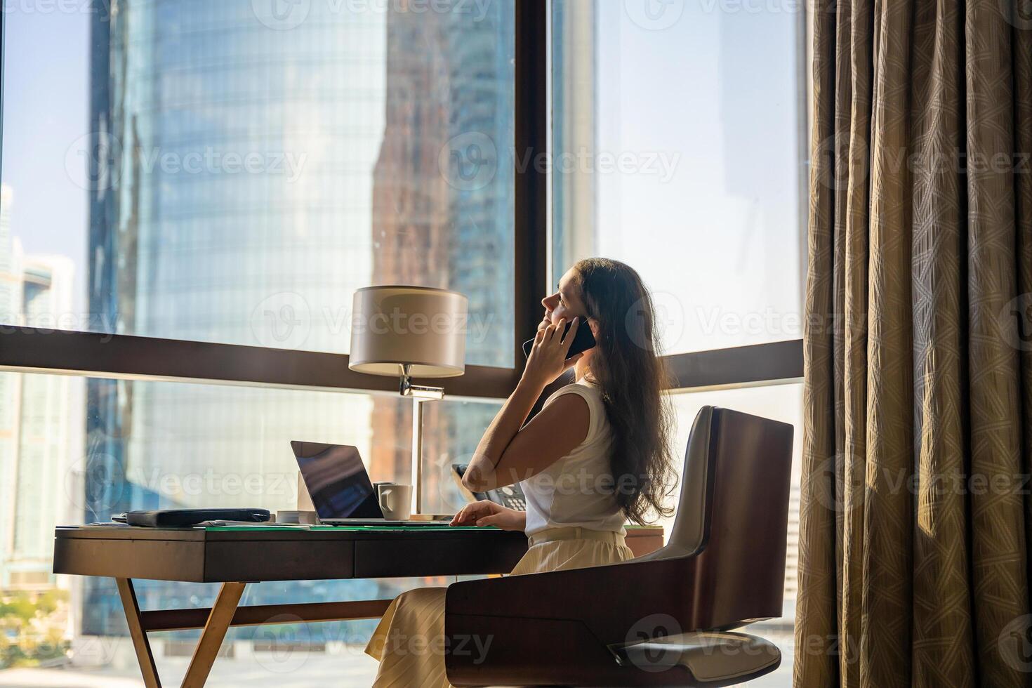 à moda empresária trabalhador autonomo trabalhando com computador portátil e inteligente telefone, ela é sentado em a cadeira, desfrutando panorâmico Visão dentro janela com cidade fundo. baixo chave foto. Alto qualidade foto