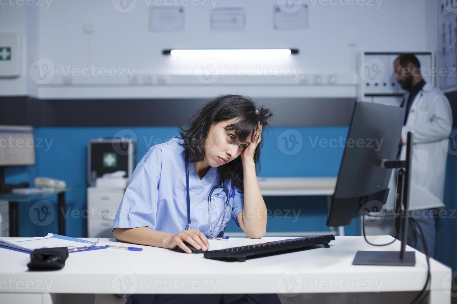 caucasiano mulher dentro azul esfoliantes, cansado estudando médico dados em dela Área de Trabalho computador. dentro uma hospital, cansado olhando fêmea enfermeira usa uma computador enquanto homem dentro uma laboratório casaco trabalho dentro fundo. foto