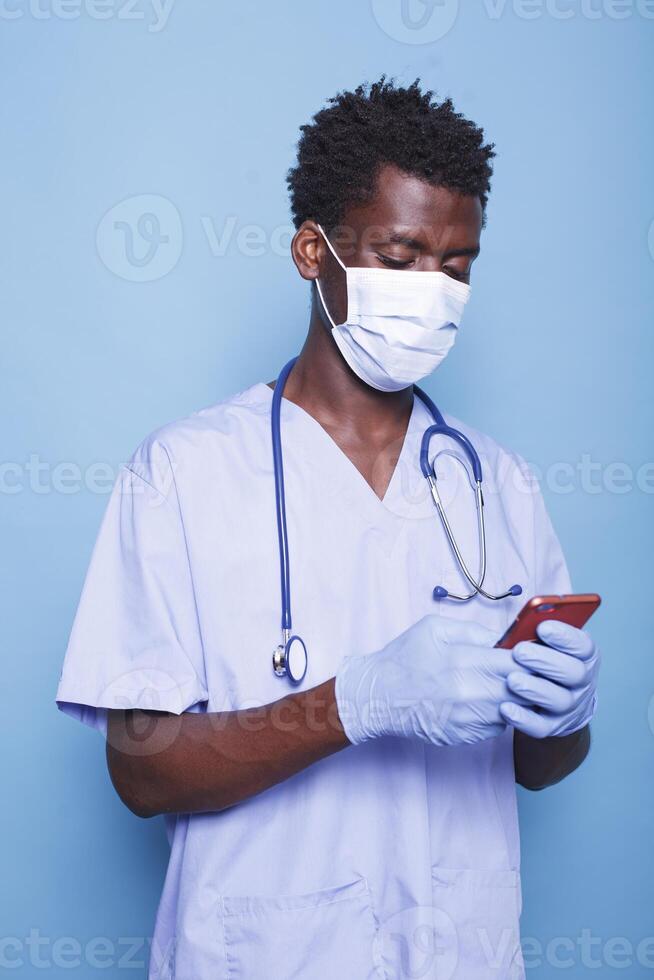 Preto homem dentro azul esfoliantes, perfil visualizar, usando uma célula telefone para médico dados e proteção durante pandemia. enfermeira digitando em Móvel dispositivo para digital cuidados de saúde enquanto em pé dentro estúdio atirar. foto