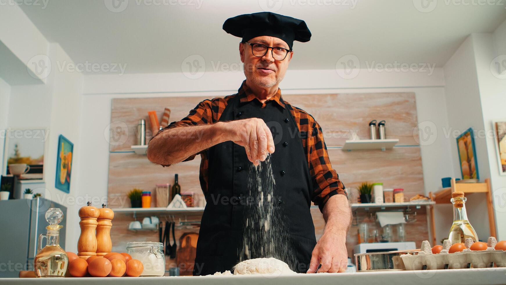 Senior homem adicionando farinha em massa de mão olhando às Câmera sorridente. aposentado idosos chefe de cozinha com bonete e uniforme aspersão, peneiramento espalhando rew ingredientes com mão cozimento caseiro pizza e pão foto