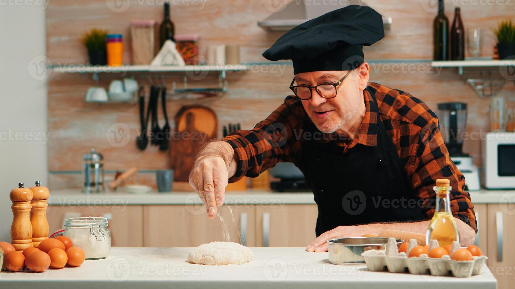 padaria homem peneiramento farinha sobre massa em mesa dentro casa cozinha. aposentado idosos chefe de cozinha com bonete e uniforme aspersão, peneirar, espalhando rew ingredientes com mão cozimento caseiro pizza e pão. foto