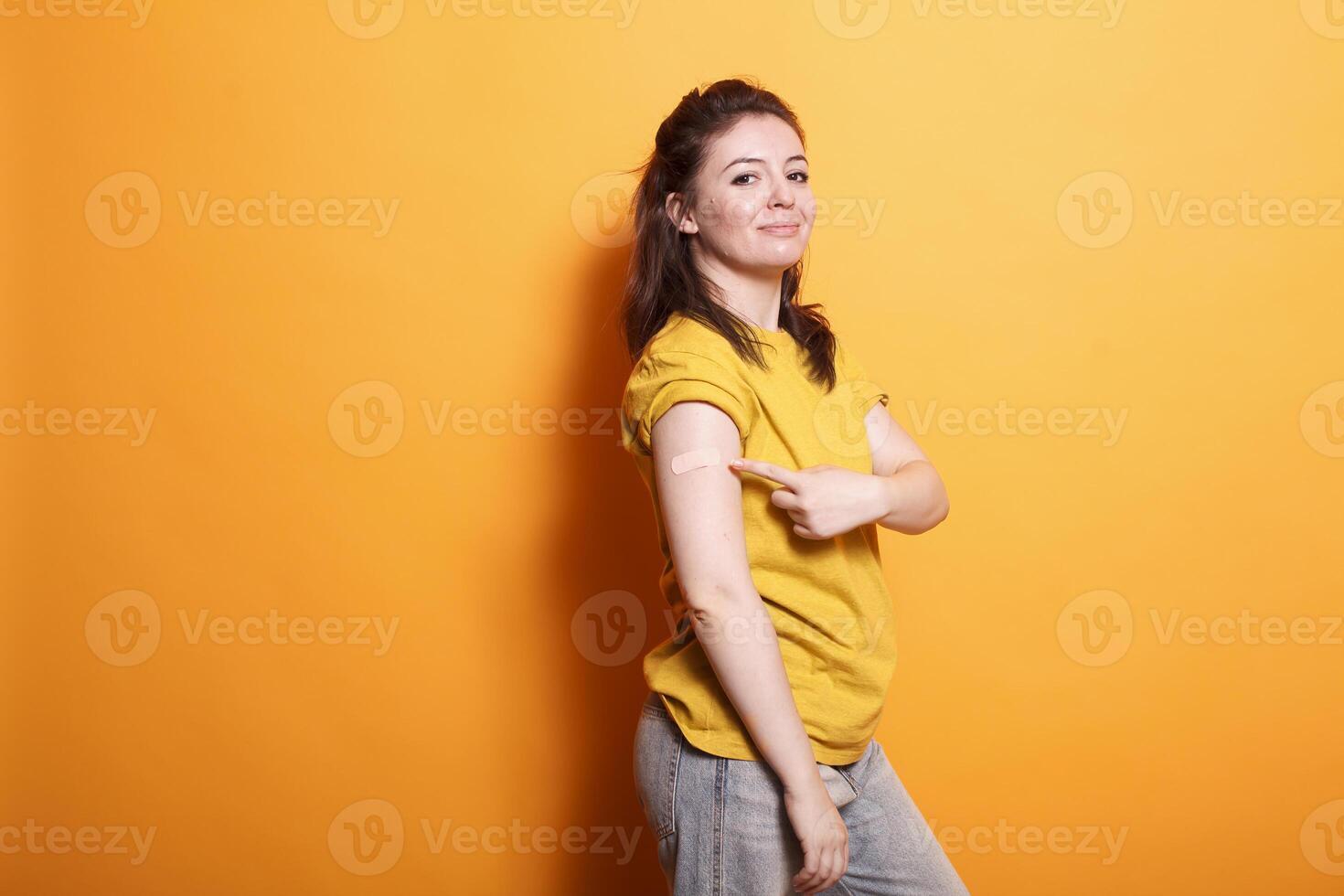 imagem mostrando uma caucasiano mulher dentro casual roupas apontando para dela braço tendo uma banda ajuda depois de uma vacinação tomada. morena senhora em pé e gesticulando para curativo a partir de imunização injeção. foto