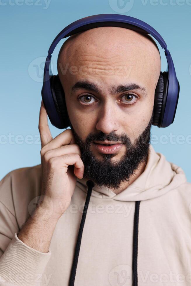 jovem árabe homem ouvindo para música dentro sem fio fones de ouvido retrato. Careca barbudo pessoa desfrutando melodias lista de reprodução dentro fones de ouvido enquanto olhando às Câmera com neutro expressão foto