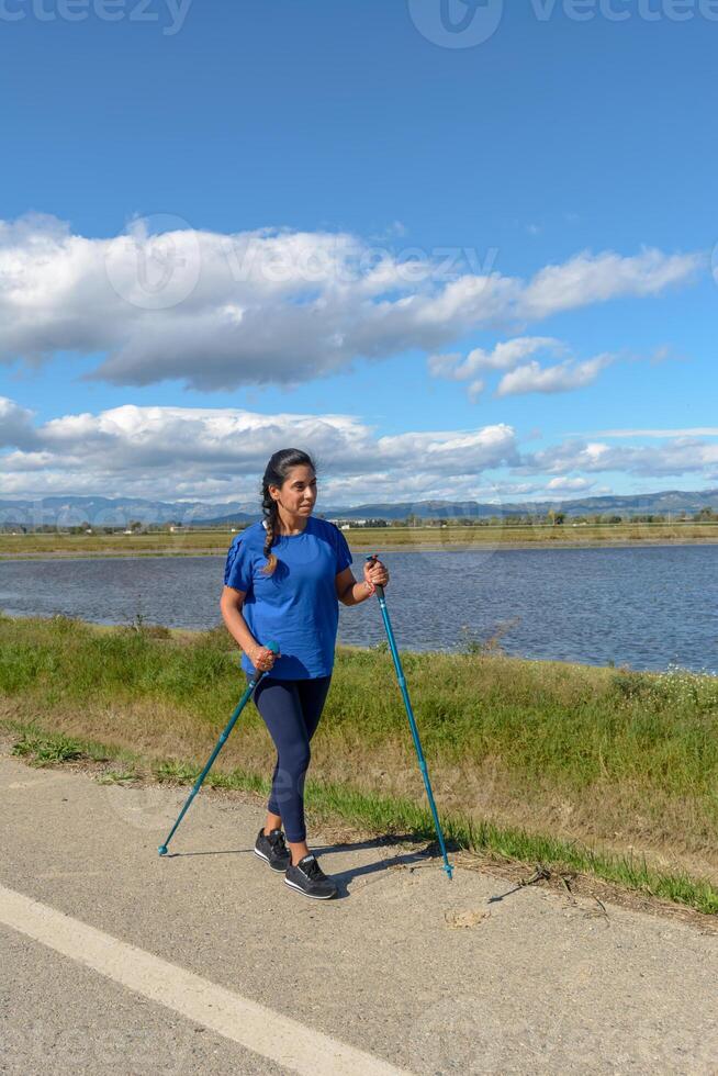 ao ar livre entusiasta com caminhando postes ao lado uma tranquilo água corpo em uma brilhante dia, hispânico latina mulher caminhando com caminhada postes dentro a Ebro delta natural parque, Tarragona, catalunha, Espanha foto