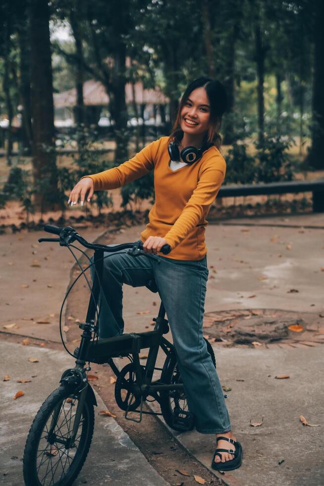 feliz jovem ásia mulher enquanto equitação uma bicicleta dentro uma cidade parque. ela sorriu usando a bicicleta do transporte. ambientalmente amigáveis conceito. foto