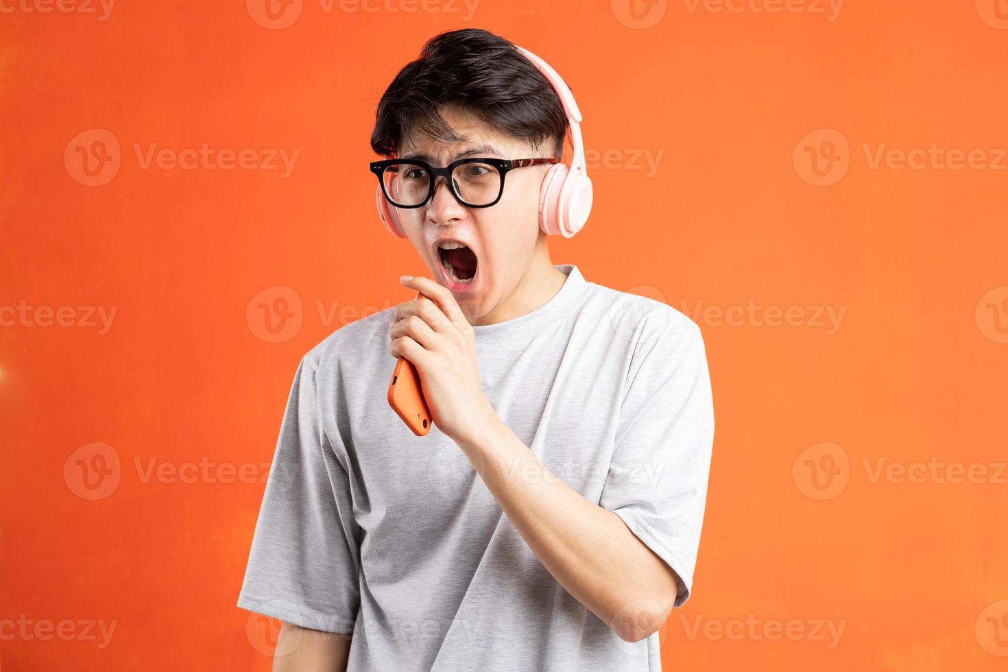 retrato de jovem asiático segurando um telefone inteligente na mão, usando fones de ouvido e cantando, isolado em um fundo laranja foto