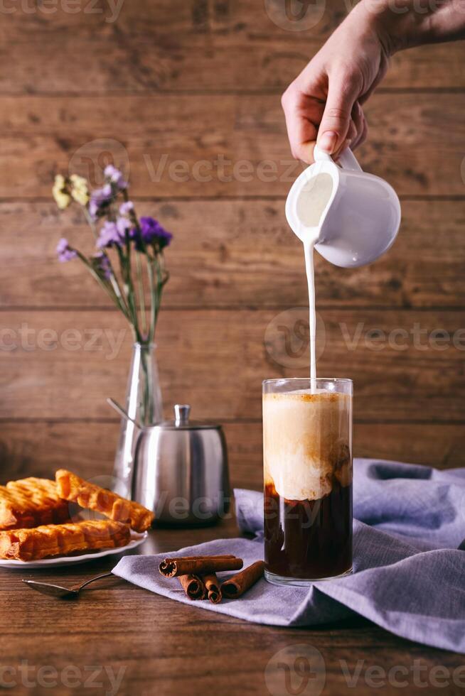mulheres mão com desnatadeira derramando leite dentro vidro com café. canela Gravetos, caseiro biscoitos e grupo do flores silvestres em uma de madeira fundo. café da manhã conceito. foto