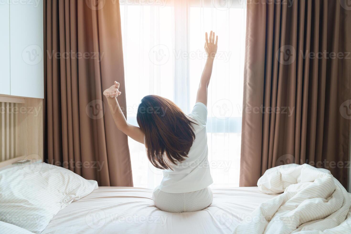 feliz mulher alongamento em cama depois de despertar acima, jovem adulto fêmea Aumentar braços e olhando para janela dentro a cedo manhã. fresco relaxar e ter uma agradável dia conceitos foto
