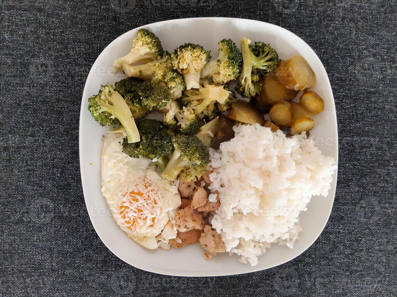 caseiro prato com brócolis, frito ovos com queijo, frango cozido carne, arroz e picles servido em branco prato foto