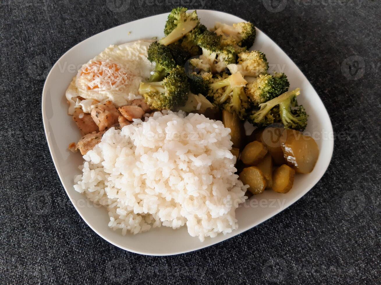 caseiro prato com brócolis, frito ovos com queijo, frango cozido carne, arroz e picles servido em branco prato foto