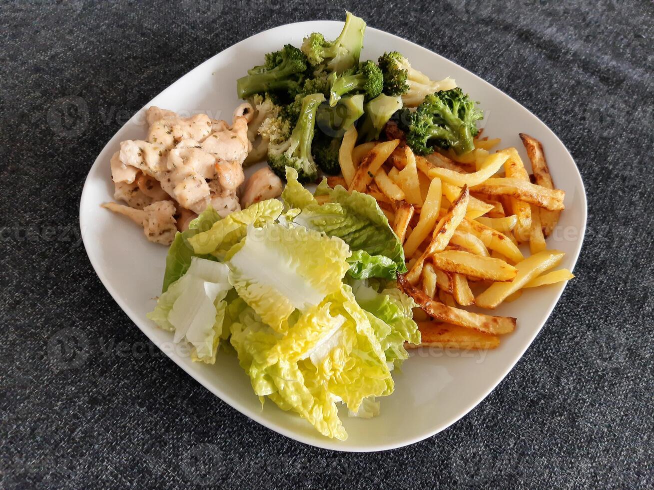 caseiro grelhado frango com francês fritas, brócolis e verde salada, servido em uma branco prato foto