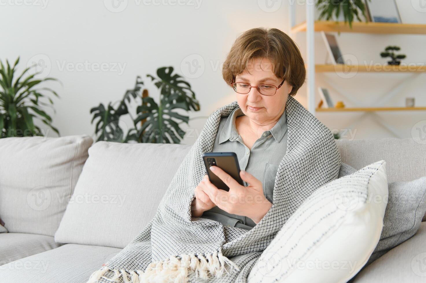 Mais velho maduro adulto mulher usando Smartphone sentado em sofá às lar. alegre avó com Óculos surfar Internet com dela Móvel telefone. tecnologia conceito foto